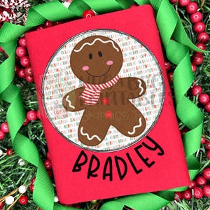 May include: A red t-shirt featuring a gingerbread man design. The gingerbread man wears a red and white striped scarf and is set within a circular design with festive greetings. The name "Bradley" is printed in black.