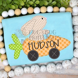 May include: A light blue t-shirt with an embroidered design of a brown bunny driving a carrot-shaped car. The car has black wheels and a checkered orange and white pattern. The bunny has brown polka dots and a pink nose. The car has the name "HUDSON" embroidered on it.