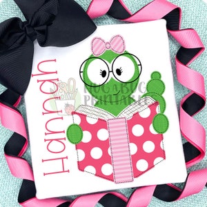 Op de afbeelding: Een groene kikker met een roze bril en een roze strik leest een roze en wit gestippeld boek. Het boek heeft een roze en wit gestreepte rug. De tekst "Happy" is in roze op een witte achtergrond geschreven.