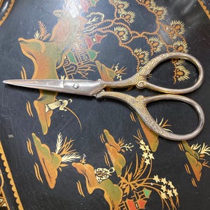 May include: A pair of silver-toned metal scissors featuring ornate, textured detailing on the handles and a sharp, pointed blade. The scissors have a classic design with two finger loops and a central screw mechanism connecting the two blades.