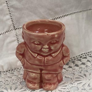 Può includere: Un portauovo vintage in ceramica a forma di ragazzo sorridente. Il portauovo è di un colore marrone-rossastro tenue, con dettagli come un colletto e una giacca abbottonata. Le mani del ragazzo sono posizionate ai lati.