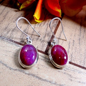 Könnte beinhalten: Ein Paar silberner Ohrhänger. Jeder Ohrring hat einen tiefmagentafarbenen, ovalen Edelstein, der in eine polierte Silberfassung gefasst ist. Die Ohrringe haben ein einfaches Haken-Design.
