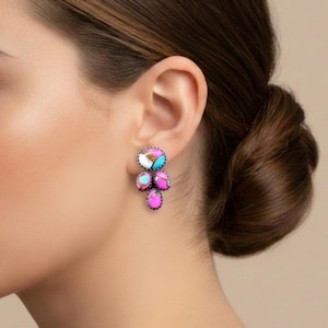 Może przedstawiać: Zbliżenie ucha kobiety z kolczykami typu cluster. Kolczyki charakteryzują się połączeniem różowych, niebieskich i białych opalizujących kamieni, osadzonych w ciemnej metalowej oprawie. Kamienie ułożone są w kaskadowy wzór.