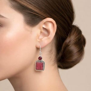 Könnte beinhalten: Silberne Ohrhänger mit einem quadratischen, tiefroten Edelstein und einem kleineren, runden roten Edelstein darüber. Die Ohrringe haben einen strukturierten Silberrand und einen einfachen Hakenverschluss.