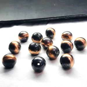 Pode incluir: Uma coleção de contas esféricas com acabamento metálico. As contas têm uma cor de base cinza escuro com detalhes em cobre. Cada conta tem um pequeno orifício, sugerindo que são para enfiar. As contas estão espalhadas sobre uma superfície branca.
