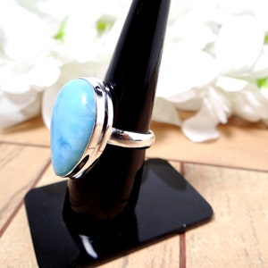 May include: A silver ring with a light blue, pear-shaped gemstone. The ring is on a black ring holder.