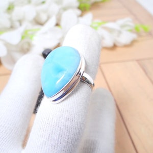 May include: A silver ring with a large, teardrop-shaped blue gemstone. The ring is set on a white fabric background.