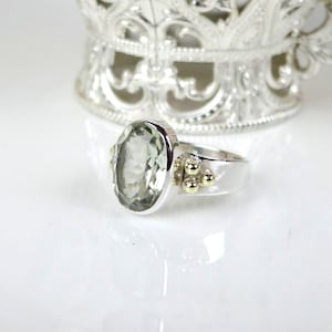 May include: A silver ring with a large, oval-cut green gemstone. The ring has gold accents and a polished finish. The gemstone is the focal point, set in a silver band with small gold details. The ring is displayed on a white surface.