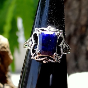 May include: A silver ring with a dark blue square gemstone set in a decorative filigree design.