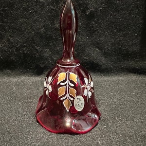 Puede incluir: Una campana de vidrio roja con un diseño floral blanco y dorado. La campana está hecha por Fenton Art Glass y tiene un borde festoneado.