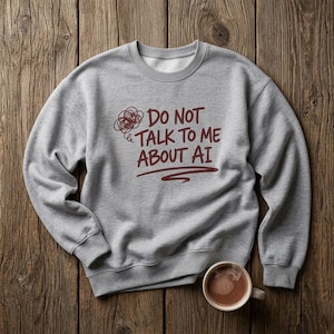 May include: A grey sweatshirt with the burgundy text "DO NOT TALK TO ME ABOUT AI". A coffee cup with a brown beverage is placed below the sweatshirt on a wooden surface. The sweatshirt has a round neck.