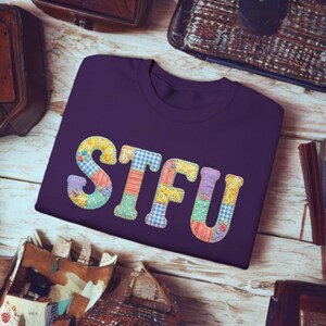 Peut inclure: Un t-shirt violet avec les lettres "STFU" appliquées dans un style patchwork. Les lettres sont composées de divers tissus à motifs, notamment des motifs floraux, vichy et rayés. Le t-shirt est présenté sur une surface en bois clair.
