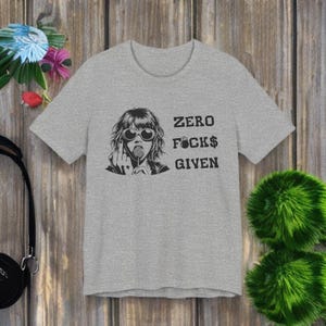 Può includere: T-shirt grigio melange con grafica di una donna con occhiali da sole e lecca-lecca, con la scritta "ZERO F*CKS GIVEN" in nero. La maglietta ha maniche corte e scollo tondo.