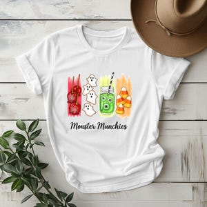Könnte beinhalten: Weißes T-Shirt mit einem Halloween-Motiv. Das Design zeigt kandierte Äpfel, Geisterkekse, ein Glas mit Augäpfeln und Zuckermais. Der Text "Monster Munchies" steht unter der farbenfrohen Illustration.