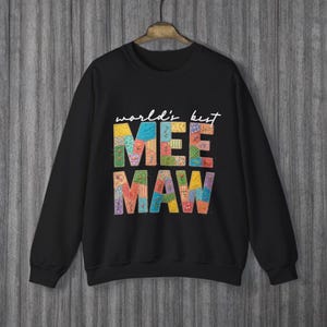 May include: Black sweatshirt with the words "world's best MEE MAW" in white script and colorful patchwork letters. The sweatshirt has long sleeves and a crew neck, perfect for casual wear.