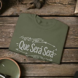 Könnte beinhalten: Olivgrünes T-Shirt mit dem Schriftzug "Que Será Será" in weißer Schrift. Der Text ist von dekorativen Pfeilen und einem Banner umgeben. Das T-Shirt liegt auf einer Holzoberfläche gefaltet.