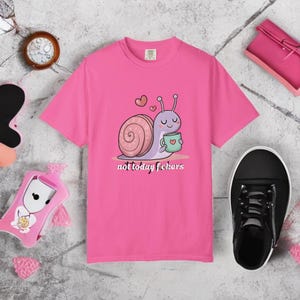 May include: A bright pink t-shirt featuring a cartoon snail holding a mug with a heart design. The snail has a light purple body and a pink shell. The text "not today fckers" is printed below the snail.
