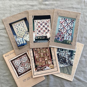 May include: A selection of vintage quilt pattern books, including designs such as "Spring Baskets," "Cottage Garden," and "Golly Gosh!" The covers display colourful quilt images with decorative borders and text. The books are arranged on a light grey fabric surface.