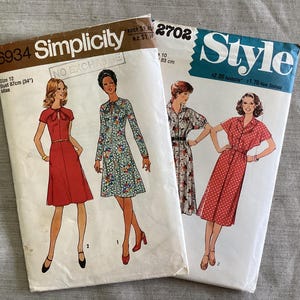 Op de afbeelding: Twee vintage naaipatronen. Eén is gelabeld "Simplicity 6934" met een rode jurk illustratie, borstomvang 87cm. De andere is "Style 2702" met een rode polkadot jurk illustratie, borstomvang 83 cm.