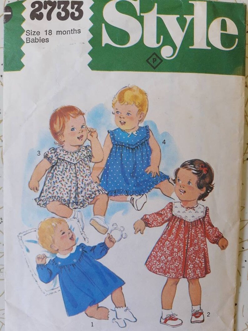 May include: Vintage sewing pattern featuring illustrations of four baby dresses. The dresses are in various styles, including a blue polka-dot dress, a red floral dress, and two dresses with floral patterns. The pattern is for size 18 months.