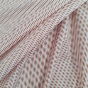 Könnte beinhalten: Weißer Stoff mit dünnen, orangefarbenen Streifen. Der Stoff ist gefaltet und hat eine strukturierte Oberfläche.