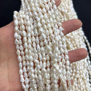 Op de afbeelding: Strengen crèmewitte zoetwaterparels. De parels zijn ovaal van vorm en hebben een glanzende glans. De afbeelding toont meerdere strengen, wat suggereert dat ze klaar zijn voor het maken van sieraden of wederverkoop. De parels worden in een hand gehouden, wat hun grootte en textuur benadrukt.