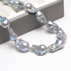 Op de afbeelding: Een ketting met iriserende barokparels. De parels zijn een mix van zilver en grijs, met hints van blauw en groen. De ketting wordt gepresenteerd op een witte ondergrond, met een grijze sieradendoos op de achtergrond.