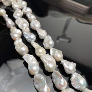 May include: Close-up of a strand of white baroque pearls. The pearls have an irregular, organic shape and a lustrous surface. The pearls are strung together, creating a necklace or bracelet.