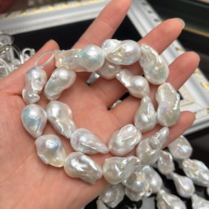 May include: A strand of large, irregularly shaped, white baroque pearls. The pearls have a lustrous, iridescent sheen, with hints of blue and green. The pearls are held in a hand, showcasing their size and unique forms.