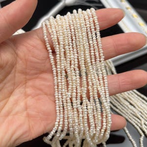 May include: Strands of small, off-white freshwater pearls. The pearls are strung together, creating multiple lines of delicate beads. The pearls have a slightly irregular shape and a subtle luster. The background is blurred, focusing attention on the pearls.