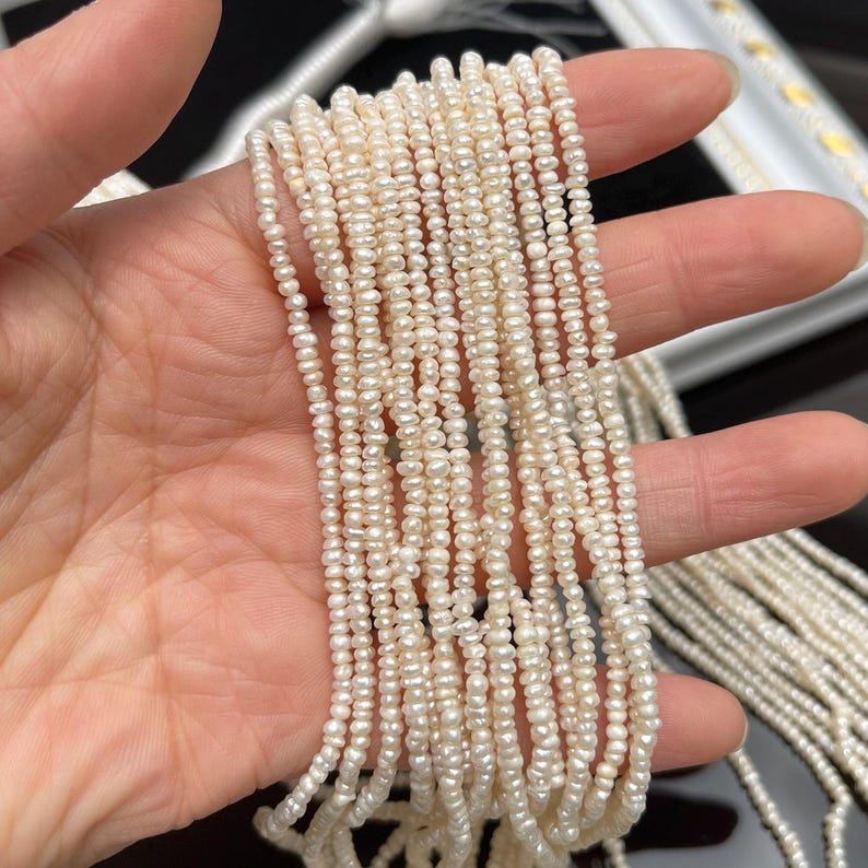 May include: Strands of small, off-white freshwater pearls. The pearls are strung together, creating multiple lines of delicate beads. The pearls have a slightly irregular shape and a subtle luster. The background is blurred, focusing attention on the pearls.