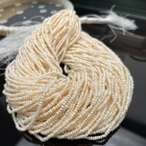 May include: A large bundle of off-white seed beads, tightly strung together. The beads have a slightly irregular shape and a subtle sheen. The strands are tied together at the top with a white thread, suggesting they are ready for jewelry making.