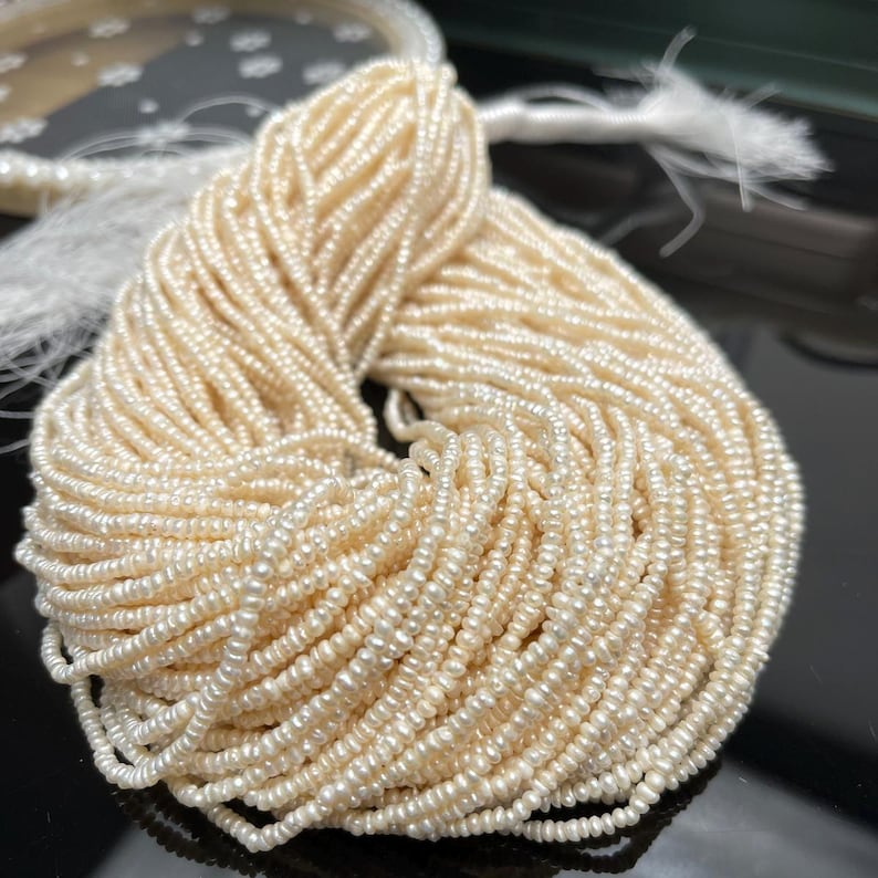 May include: A large bundle of off-white seed beads, tightly strung together. The beads have a slightly irregular shape and a subtle sheen. The strands are tied together at the top with a white thread, suggesting they are ready for jewelry making.