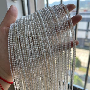 Puede incluir: Un conjunto de collares de perlas blancas, sostenidos en una mano. Los hilos de perlas son numerosos y cuelgan, mostrando su delicadeza. Las perlas son pequeñas y tienen un brillo sutil, creando un aspecto suave y elegante.