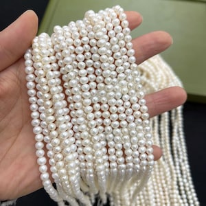 Könnte beinhalten: Nahaufnahme von mehreren Strängen kleiner, weißer Perlen. Die Perlen sind rund und haben einen glänzenden Glanz. Die Stränge werden in einer Hand gehalten, der Hintergrund ist unscharf. Die Perlen scheinen eng aneinandergereiht zu sein.