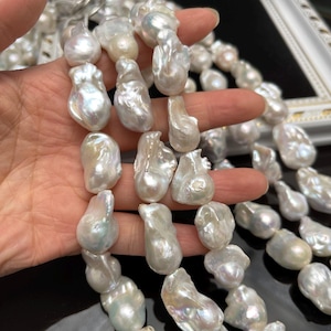 May include: A strand of large, irregular white baroque pearls with a high-luster, iridescent finish. The pearls are strung together, showcasing varied, organic shapes and smooth, reflective surfaces that catch the light with subtle pink and blue tones.