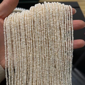 May include: Strands of small, off-white pearls, tightly strung together. The pearls have a slightly irregular shape, creating a textured surface. The pearls are held in a hand, with a dark background.