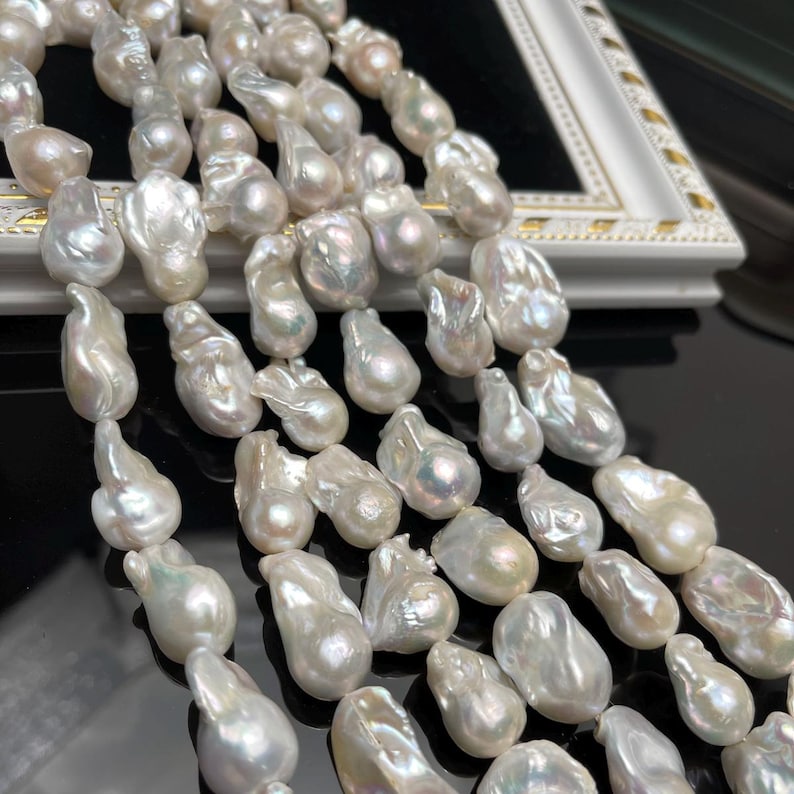 May include: Close-up of multiple strands of lustrous, irregularly shaped white pearls. The pearls have a natural, organic form, with a creamy white color and subtle iridescent sheen. The pearls are displayed against a white and gold framed surface.