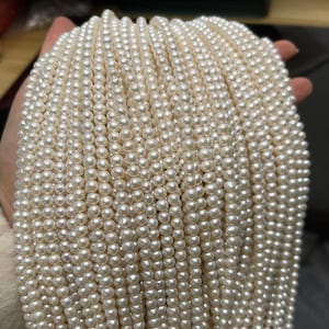 Puede incluir: Una colección de hilos de perlas blancas cremosas. Las perlas son pequeñas, redondas y de tamaño uniforme, creando una superficie lisa y brillante. Los hilos se mantienen unidos, mostrando la delicada naturaleza de la joyería.