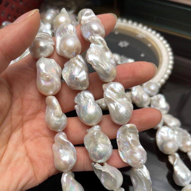 May include: A strand of large, irregularly shaped, lustrous white pearls. The pearls have a creamy, iridescent sheen, with a variety of organic forms. A white pearl necklace is visible in the background.
