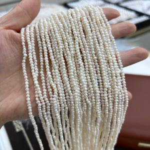 May include: A collection of multiple strands of small, off-white, irregular seed pearls. The pearls are strung together in long, vertical lines, showcasing a natural, textured surface and a soft, lustrous finish across each individual bead.