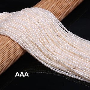 Pode incluir: Fios de pérolas ovais brancas cremosas. As pérolas são amarradas, criando um efeito de cascata. As pérolas estão sobre uma superfície de madeira. O texto "AAA" é visível no canto inferior esquerdo.