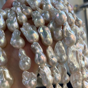 May include: Close-up of a strand of lustrous, irregularly shaped baroque pearls. The pearls have a creamy white color with iridescent overtones, reflecting light. The pearls are strung together, creating a necklace or bracelet.
