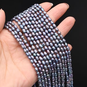 Könnte beinhalten: Stränge dunkelgrauer, oval geformter Perlen. Die Perlen haben einen subtilen, irisierenden Glanz mit violetten und blauen Farbtönen. Die Perlen werden in einer Hand vor schwarzem Hintergrund gehalten. Die Perlen sind aufgefädelt.