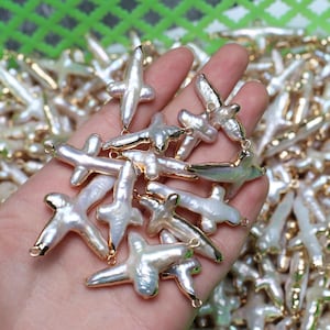 Può includere: Una collezione di pendenti a forma di croce in perle bianche con dettagli in metallo color oro. Le perle hanno una forma irregolare e organica. I pendenti sono tenuti in una mano, con altri visibili sullo sfondo, suggerendo un tema di gioielleria.