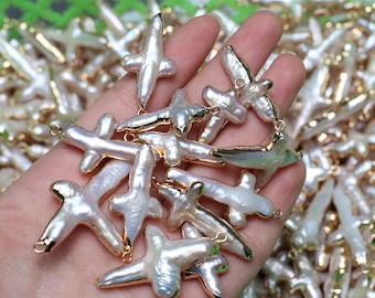 May include: A collection of white, cross-shaped pearl pendants with gold-colored metal accents. The pearls have an irregular, organic shape. The pendants are held in a hand, with more visible in the background, suggesting a jewelry-making theme.