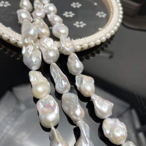 May include: A strand of irregular, baroque-shaped white pearls with a high-luster, iridescent finish. The pearls exhibit natural variations in size and shape, displaying subtle pink and blue undertones across their textured, organic surfaces.