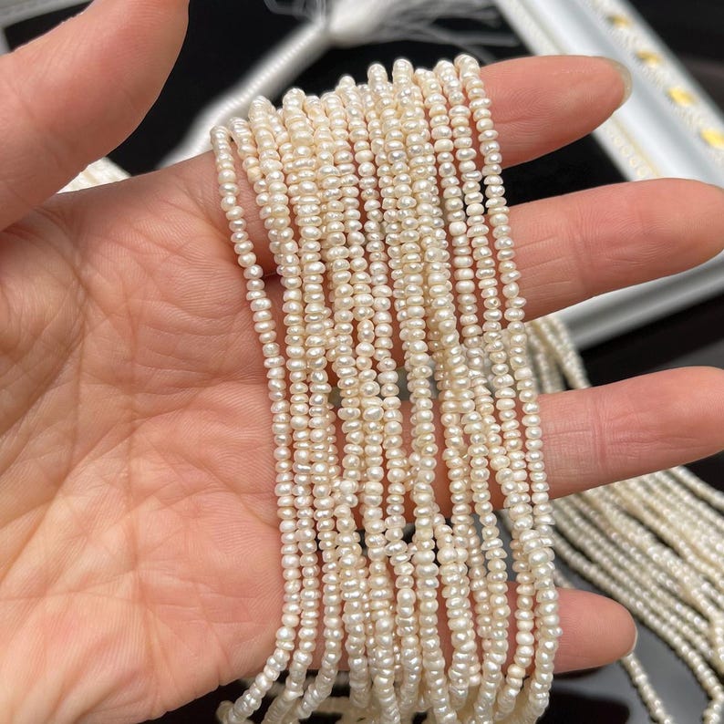 May include: A collection of delicate, off-white pearl strands. The pearls are small and irregularly shaped, creating a textured appearance. The strands are held in a hand, showcasing their length and the subtle variations in color and size.