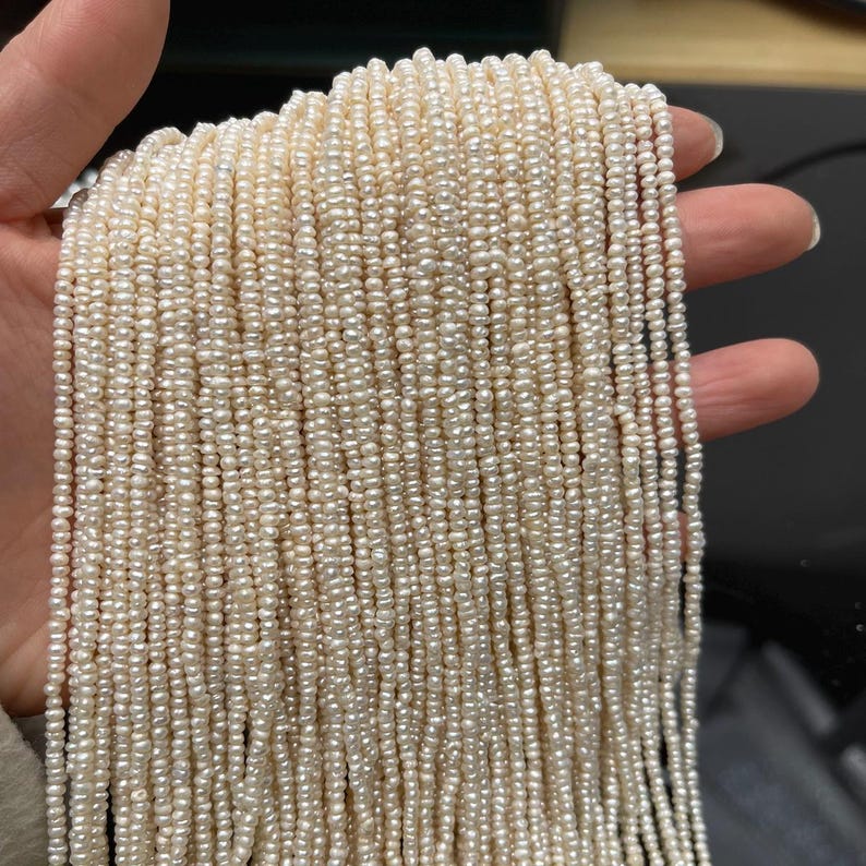 May include: A bundle of creamy white, small, irregular-shaped pearls. The pearls are strung together, creating a cascade of strands. The pearls are held in a hand, showcasing their color and texture. Ideal for jewelry making.
