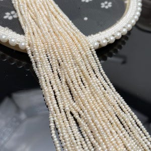 May include: A close-up shot of multiple strands of small, off-white pearls. The pearls are strung together, creating a cascading effect. The pearls are resting on a white, lace-trimmed surface with a pearl border. The background is a dark, reflective surface.
